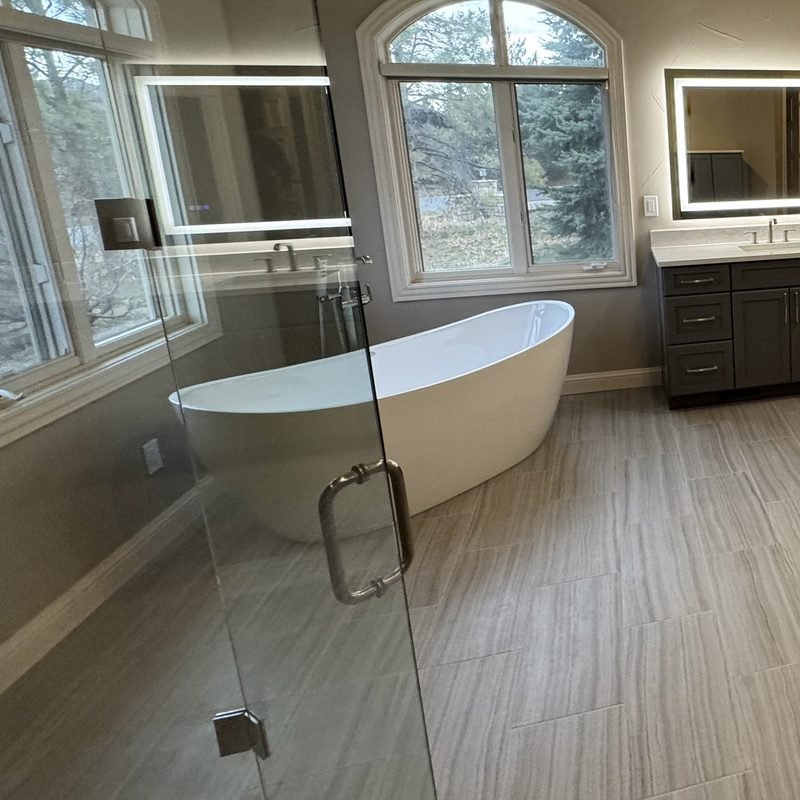 A white freestanding soaking tub under an arched window, next to a dark double vanity with lit mirrors