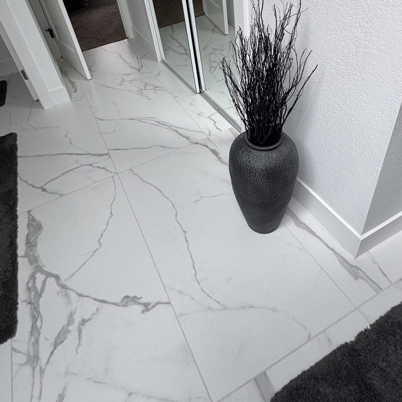 Large format white marble-look porcelain floor tile with grey veining, with a tall black vase standing where the floor meets the wall