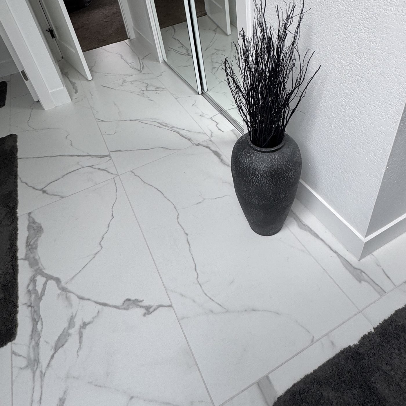 Large format white marble-look porcelain floor tile with grey veining, with a tall black vase standing where the floor meets the wall